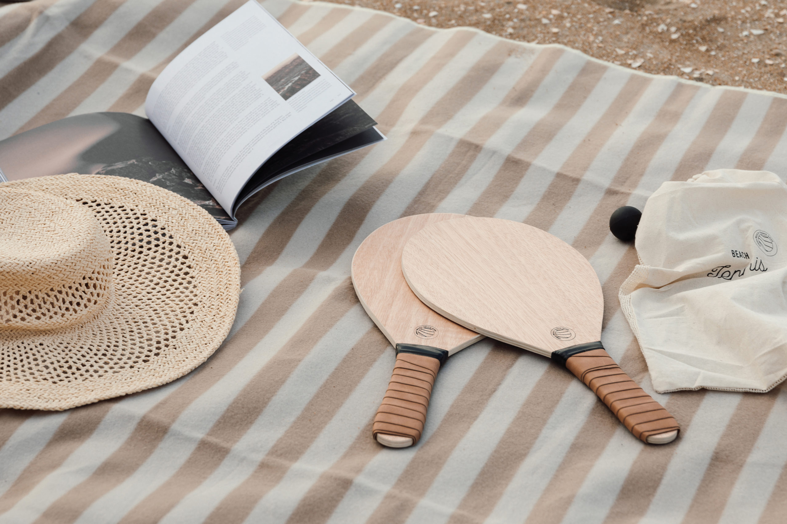 two beach rackets, a book and a hat on a beach towel